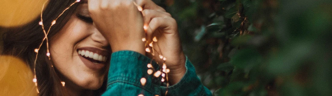 Dentist Flower Mound TX Cosmetic Dentistry Focused A person with dark hair and a bright smile is partially obscured by a string of warm-toned fairy lights held in their hand.
