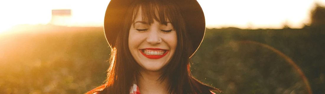 Dentist Flower Mound TX Implants Focused Bathed in warm light, a smiling woman with dark hair, bangs, and bright red lipstick wears a hat while closing her eyes.