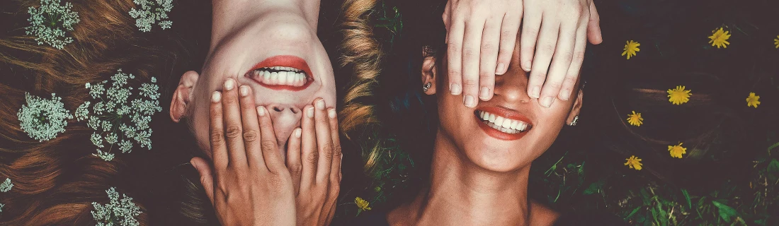 Dentist Flower Mound TX Invisalign Focused Two smiling individuals lie side-by-side on grass with white and yellow flowers with their hands over each other's eyes.